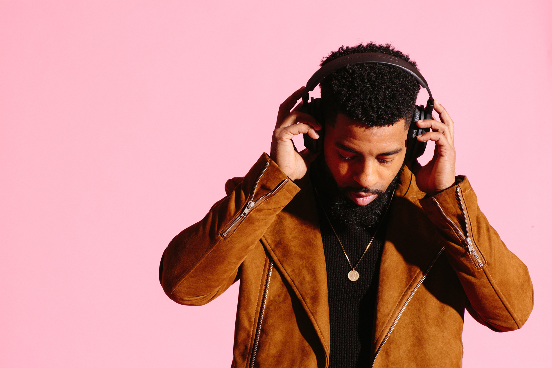 Man holding headphones to his ears standing in front of pink background.