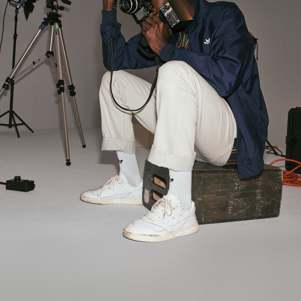 Man sat on old suitcase holding a camera and wearing a blue jacket, white trousers, white socks and white adidas sneakers.